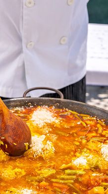 A chef stirs a large pan of vibrant vegetable paella with a wooden spoon on an outdoor table.