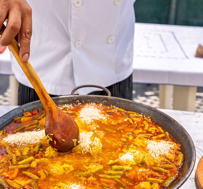 A chef stirs a large pan of vibrant vegetable paella with a wooden spoon on an outdoor table.
