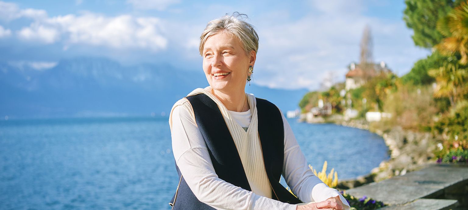 A smiling woman with short gray hair stands by a lake on a sunny day, with mountains and trees in the background.