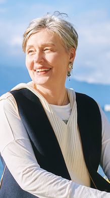 A smiling woman with short gray hair stands by a lake on a sunny day, with mountains and trees in the background.