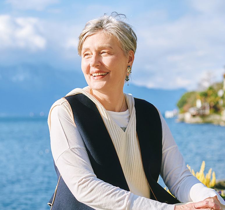 A smiling woman with short gray hair stands by a lake on a sunny day, with mountains and trees in the background.