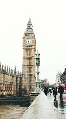 Wide view of the Houses of Parliament and Big Ben in London, with a red double-decker bus crossing Westminster Bridge on a cloudy day.