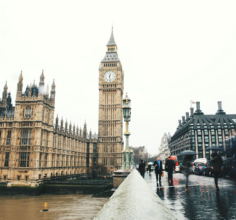 Wide view of the Houses of Parliament and Big Ben in London, with a red double-decker bus crossing Westminster Bridge on a cloudy day.