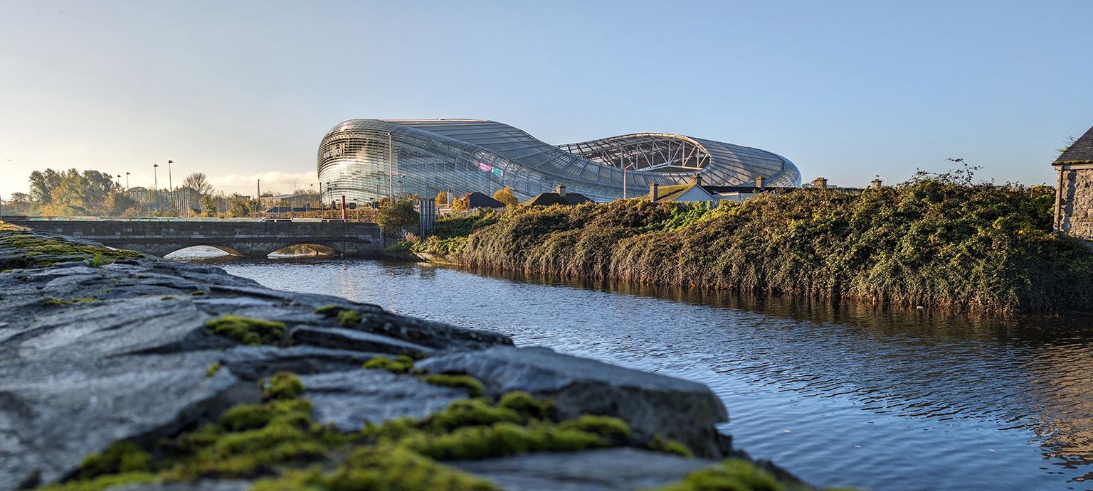 Modern stadium with a curved roof next to a river, viewed from a moss-covered stone wall, under a clear blue sky.