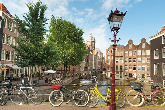A scenic canal in Amsterdam with bicycles lined up along the railing, historic buildings, trees, and a streetlamp under a blue sky.