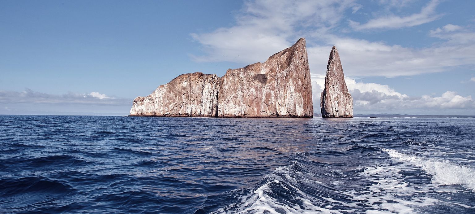 A rugged rock formation, known as Kicker Rock, rises from the ocean under a partly cloudy sky, with waves rippling in the foreground.