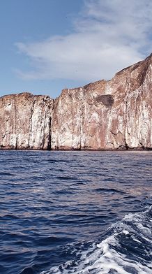 A rugged rock formation, known as Kicker Rock, rises from the ocean under a partly cloudy sky, with waves rippling in the foreground.
