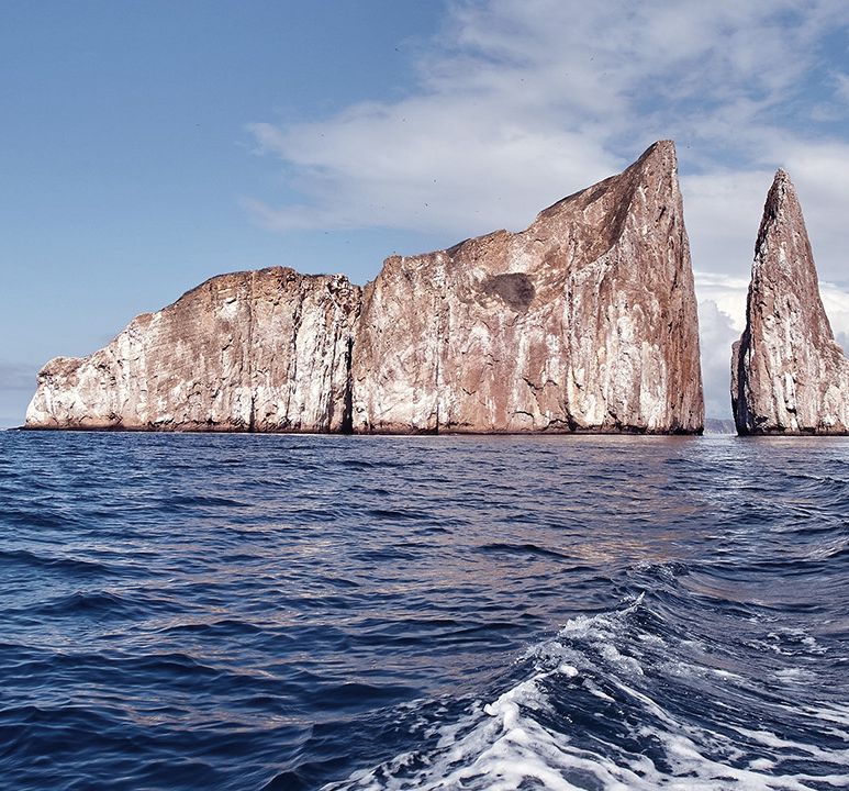 A rugged rock formation, known as Kicker Rock, rises from the ocean under a partly cloudy sky, with waves rippling in the foreground.