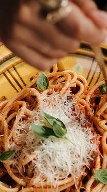 A person twirls spaghetti with tomato sauce, grated cheese, and basil using a fork on a yellow plate, over a checkered tablecloth.