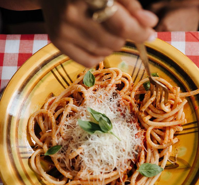 A person twirls spaghetti with tomato sauce, grated cheese, and basil using a fork on a yellow plate, over a checkered tablecloth.