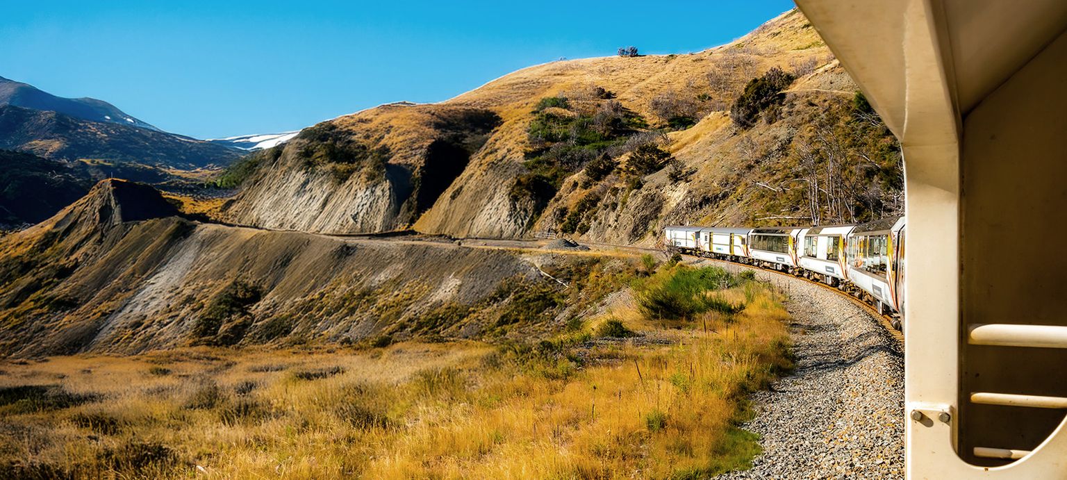 A train curves along a scenic mountain landscape with dry grass and rocky cliffs under a clear blue sky.