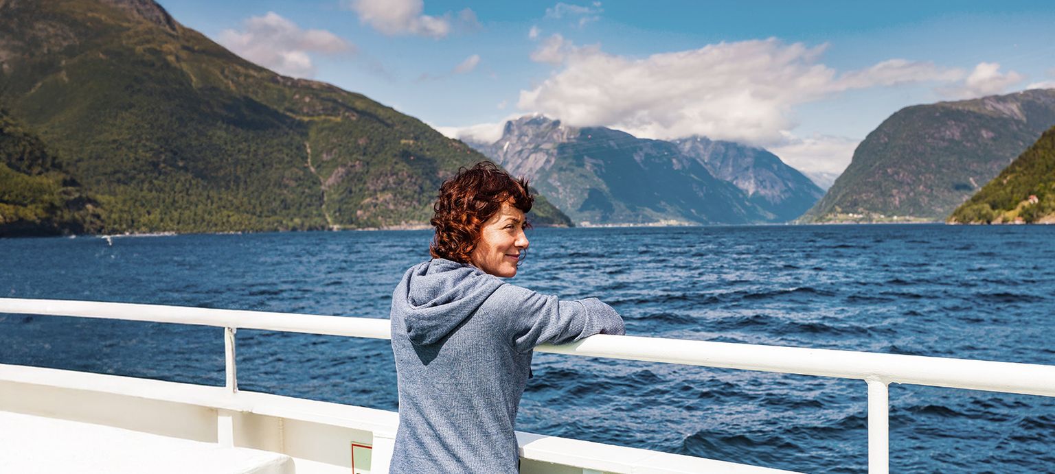 Woman in a blue hoodie leans on a ship's railing, gazing at a scenic fjord with mountains and blue sky in the background.