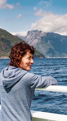 Woman in a blue hoodie leans on a ship's railing, gazing at a scenic fjord with mountains and blue sky in the background.