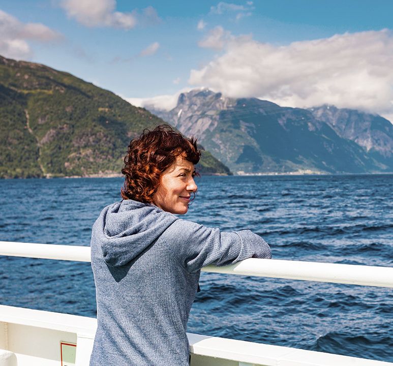 Woman in a blue hoodie leans on a ship's railing, gazing at a scenic fjord with mountains and blue sky in the background.