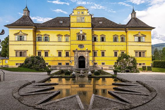 Bright yellow baroque-style building with ornate statues, a symmetrical facade, and a star-shaped fountain in the foreground, surrounded by greenery.