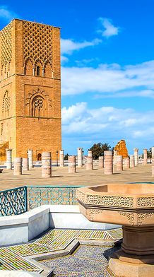 A stone tower stands amid numerous columns under a blue sky. In the foreground, a decorative fountain with colorful tiles is visible.
