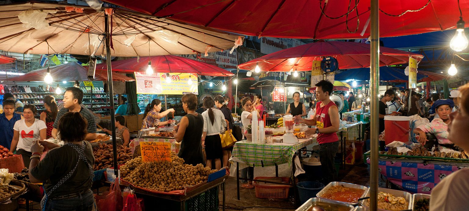 Busy night market with red umbrellas, food stalls, and diverse people shopping and selling various goods under bright hanging lights.
