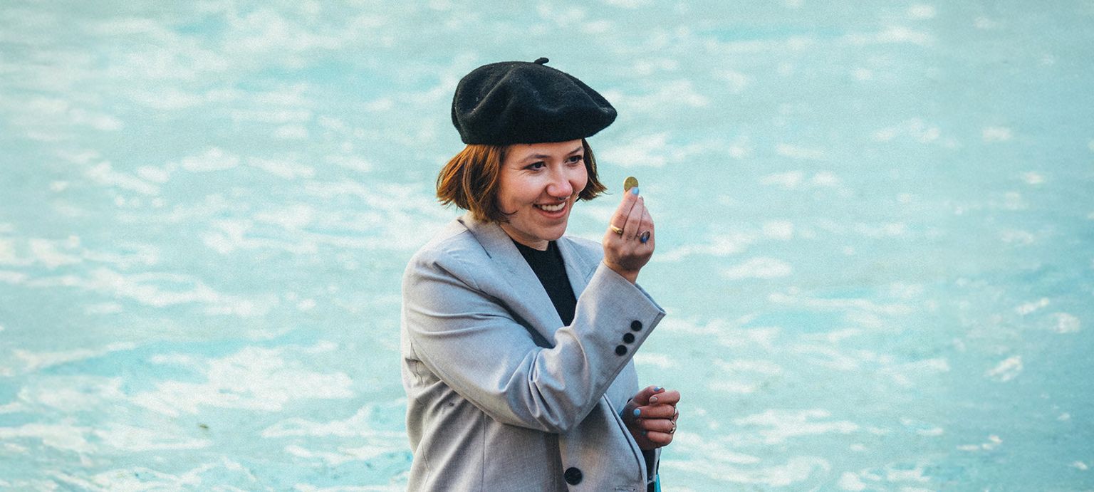 Smiling woman in a beret holds a coin near a fountain, dressed in a light coat against a blue water backdrop.