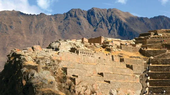Ancient stone terraces and ruins on a mountain slope with people exploring, against a backdrop of rugged brown mountains and a blue sky.