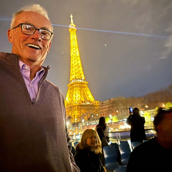 Smiling man in foreground with the illuminated Eiffel Tower in the background during the night, surrounded by seated and standing people.