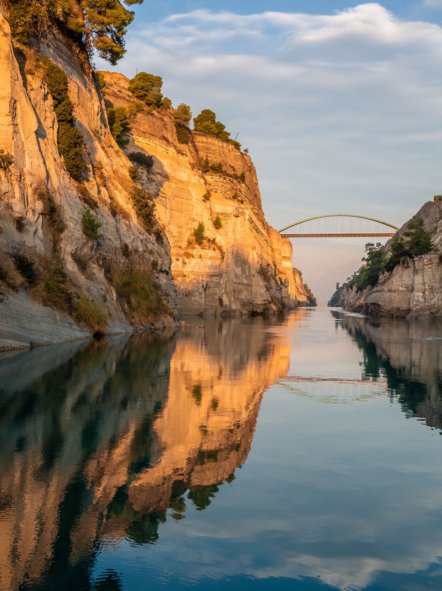 Golden cliffs reflected in calm water, with a distant arched bridge under a partly cloudy sky. Trees dot the rocky landscape.