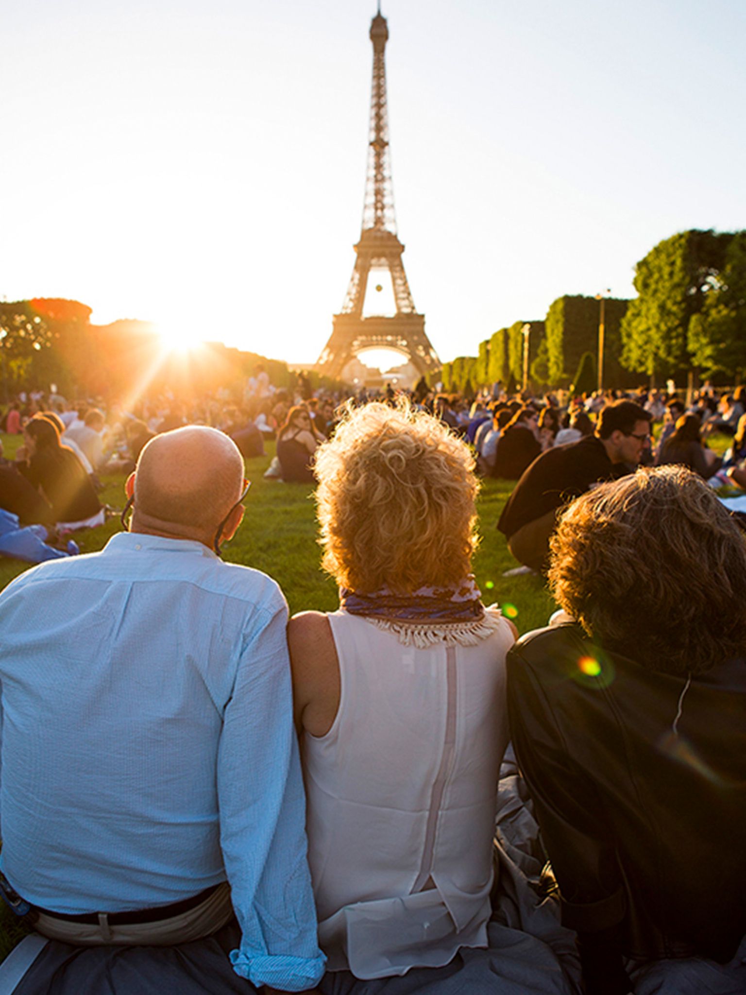 People sitting on grass at sunset, facing the Eiffel Tower. Sunlight creates a warm atmosphere, with a crowd in the background.