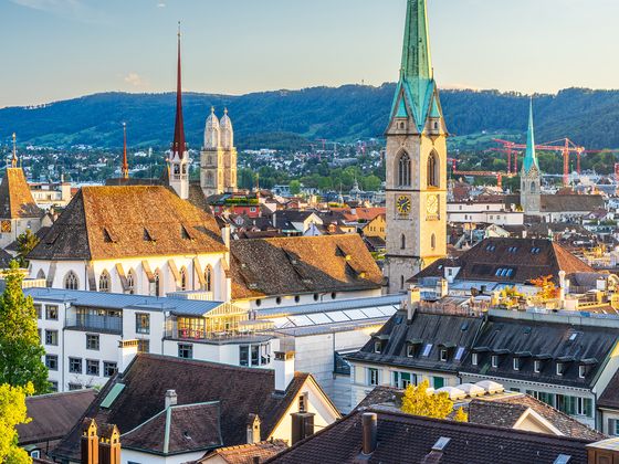 A scenic view of Zurich with historic buildings, church spires, and lush greenery under a clear blue sky.