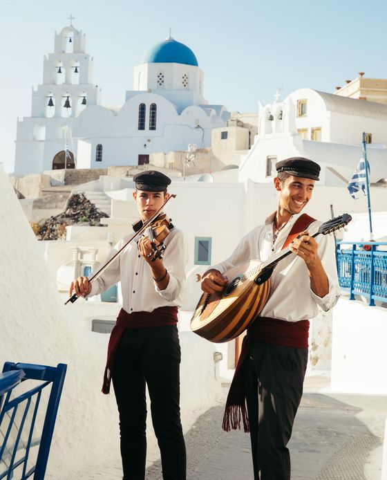 Two musicians in traditional attire play violin and guitar in a sunlit Greek village with white buildings and a blue-domed church.