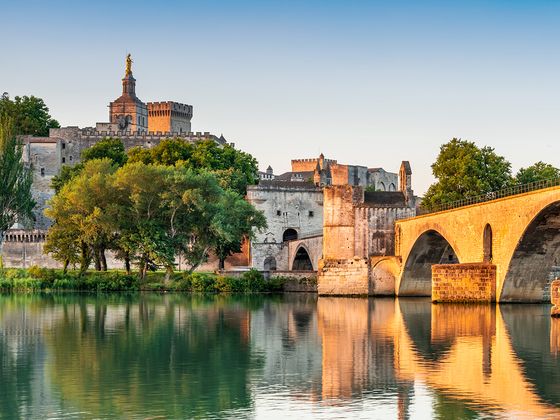 A stone bridge reflects in a calm river with an ancient building and trees in the background during sunset.