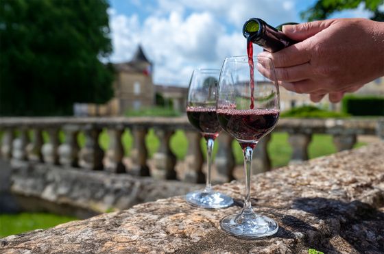 Red wine being poured into a glass on a stone balustrade with a blurred chateau and garden in the background under a partly cloudy sky.