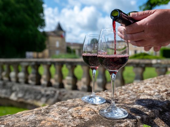 Red wine being poured into a glass on a stone balustrade with a blurred chateau and garden in the background under a partly cloudy sky.