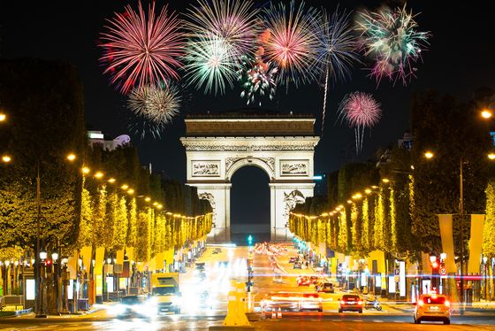 Arc de Triomphe at night with colorful fireworks in the sky, bright streetlights, and cars on the busy Champs-Élysées below.