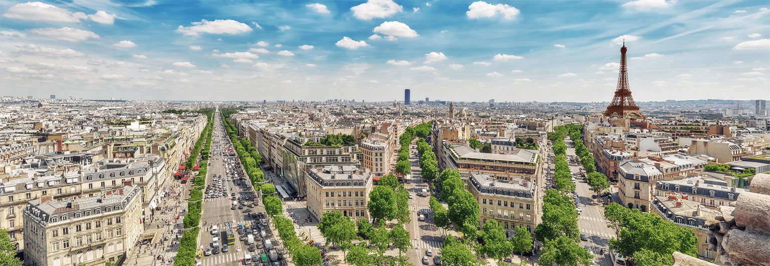 Aerial view of Paris with wide boulevards lined by trees, the Eiffel Tower, and cityscape under a bright blue sky with clouds.