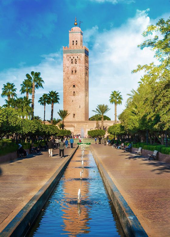 A tall, ornate tower stands amidst palm trees, with a walkway and narrow reflecting pool leading towards it, under a bright blue sky.