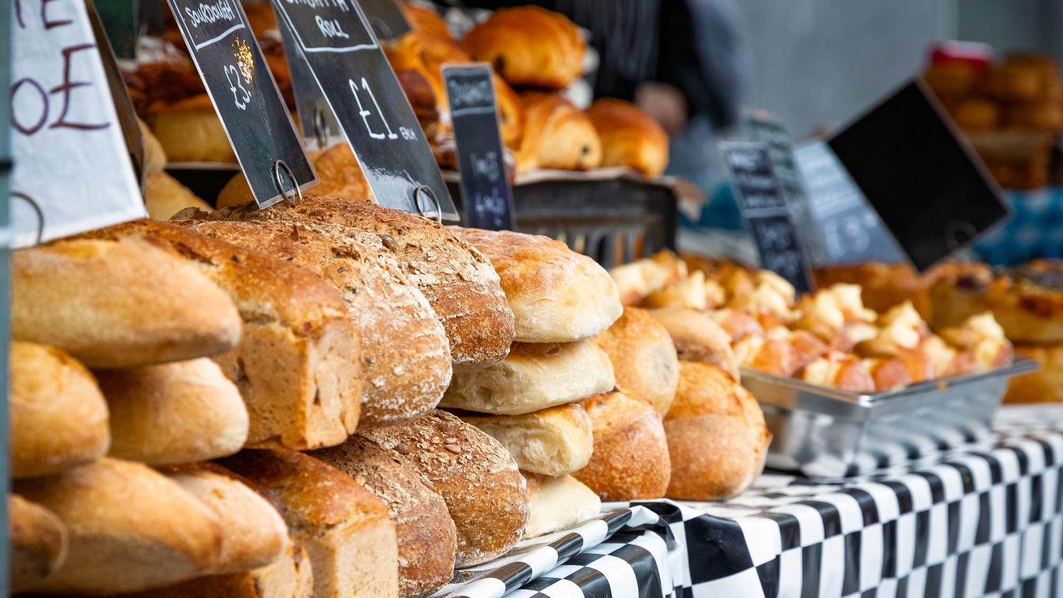 Assorted loaves and pastries displayed on a checkered tablecloth at a market, with price signs visible.