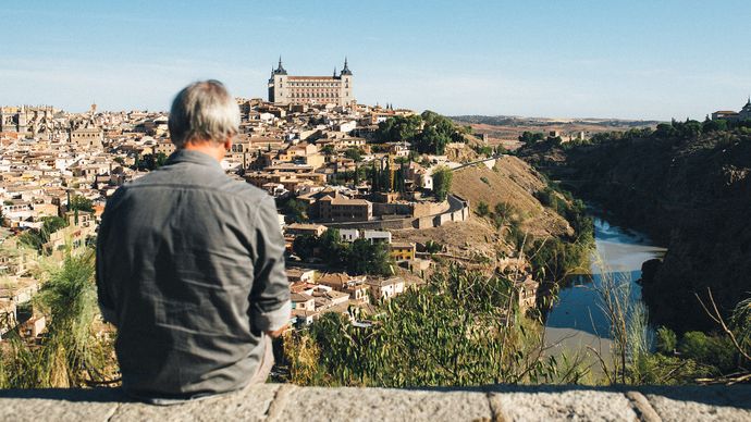 A person sits on a ledge overlooking the historic cityscape of Toledo, Spain, with the Alcázar and the Tagus River visible under a clear blue sky.
