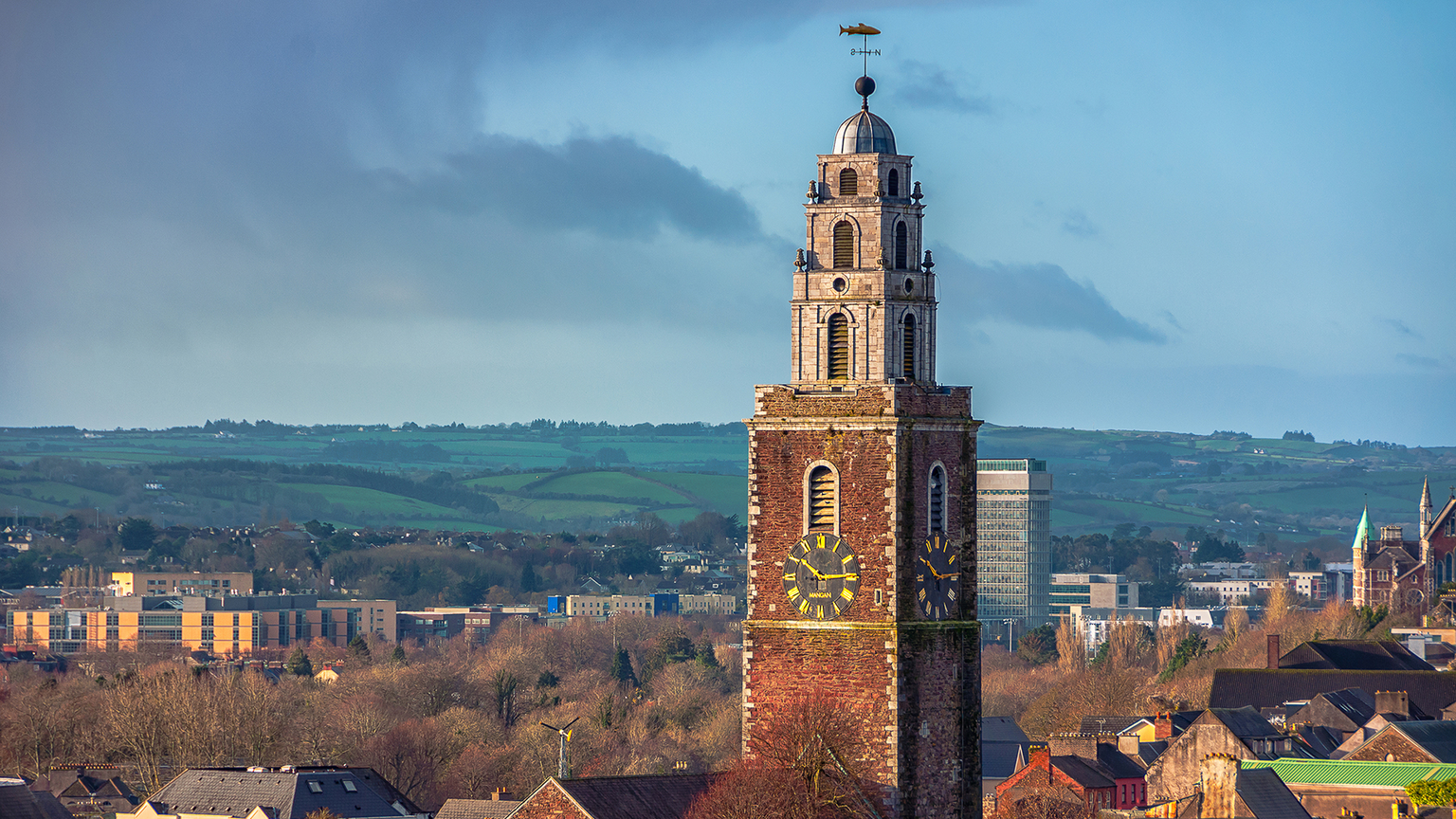 Clock tower with a weather vane against a backdrop of green hills and a cloudy blue sky.