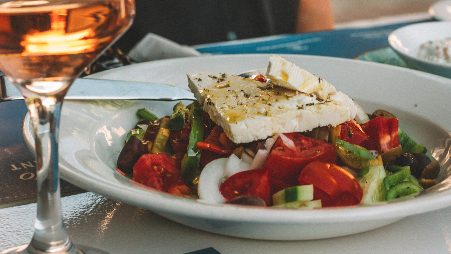 A fresh Greek salad with feta, tomatoes, cucumbers, onions, and olives on a white plate. A glass of wine is in the foreground.