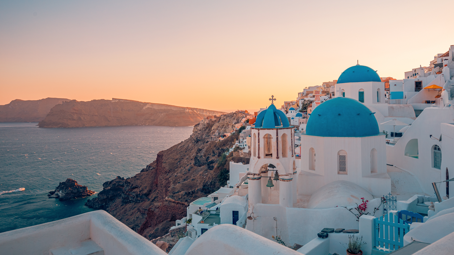 Santorini at sunset, featuring white buildings with blue domes overlooking the sea, rugged coastline, and a warm, colorful sky.