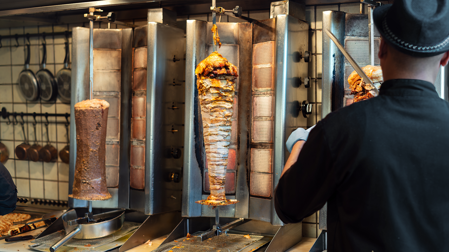 A chef slices meat off a vertical rotisserie in a kitchen with multiple meat spits, pans hanging on the tiled wall in the background.