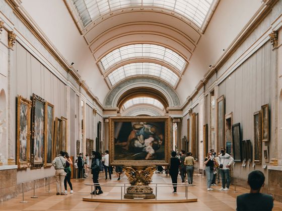 People admire artworks in a spacious, high-ceilinged gallery with ornate moldings and framed paintings lining the walls.