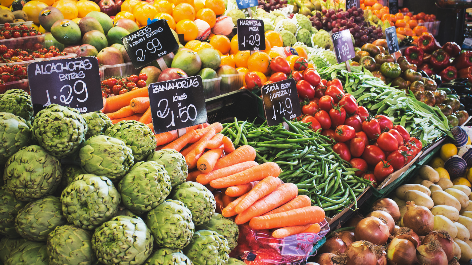 A colorful market stall displays artichokes, carrots, green beans, tomatoes, and various fruits with price tags.