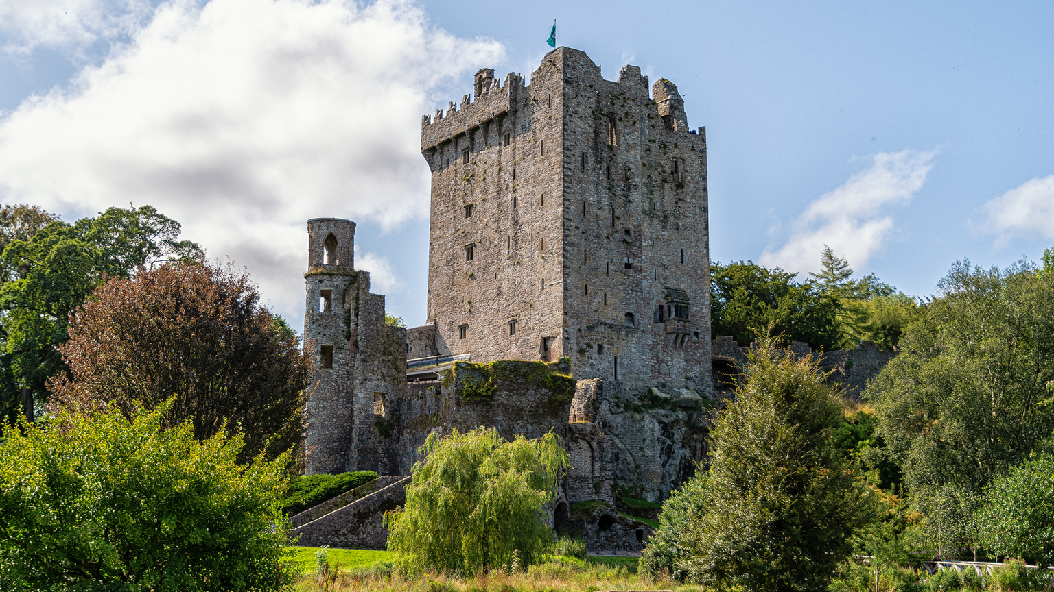 Stone castle with tall tower, surrounded by lush greenery and trees under a partly cloudy sky.