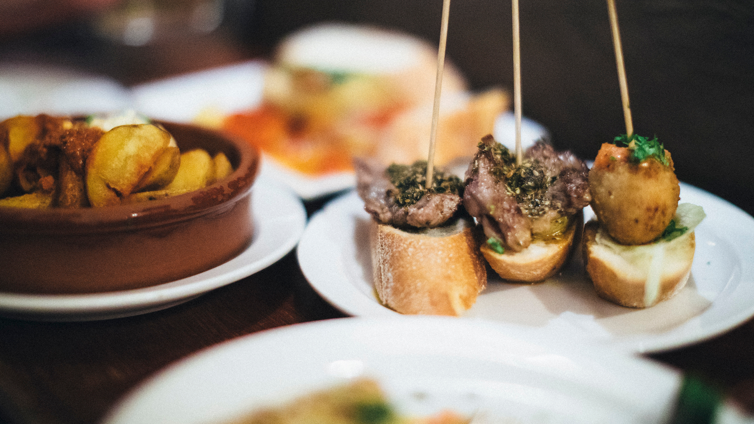 Close-up of various tapas on a table, including skewered meat on bread and a bowl of potatoes in sauce, on white plates.