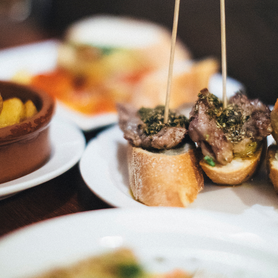 Close-up of various tapas on a table, including skewered meat on bread and a bowl of potatoes in sauce, on white plates.