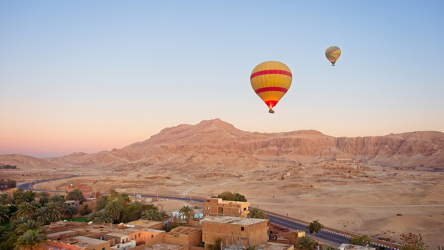 Two hot air balloons float over a desert landscape with sandy mountains and small buildings at sunset.