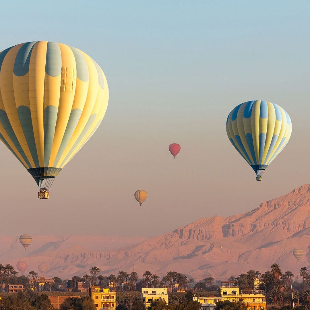 Hot air balloons in a clear sky above a desert landscape with distant mountains and scattered buildings at sunrise.