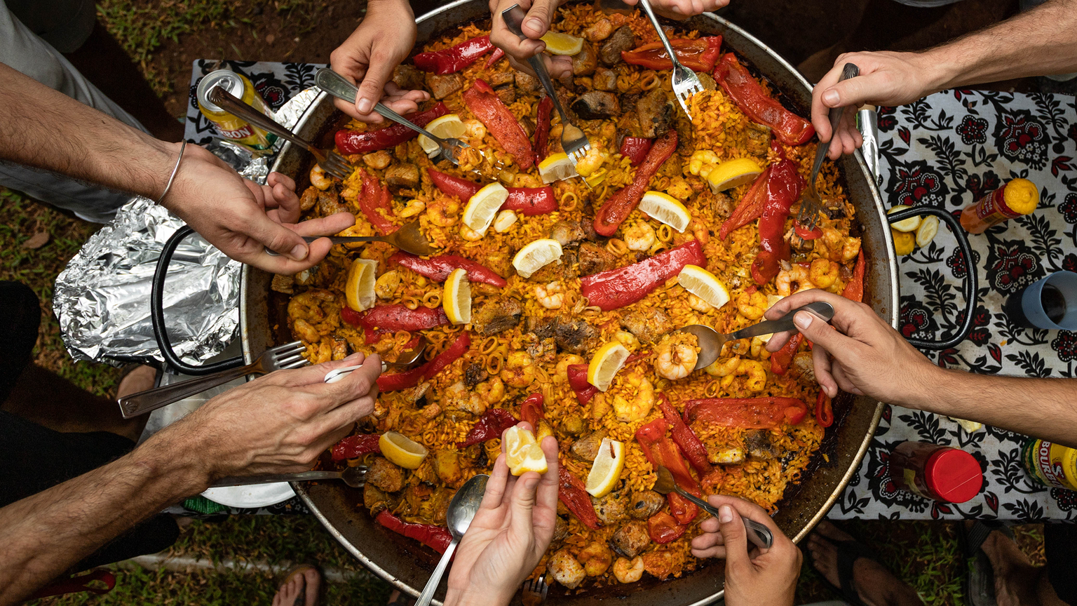 Several hands reach into a large pan of colorful paella with seafood, lemon wedges, and red peppers, set on a patterned tablecloth.