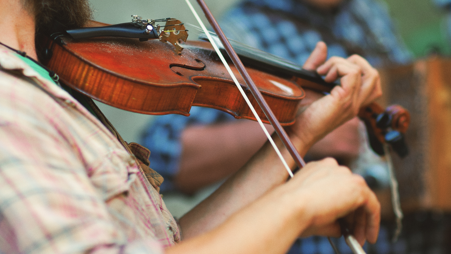 A person playing a violin with a focus on the strings and bow, wearing a plaid shirt. Another musician is blurred in the background.