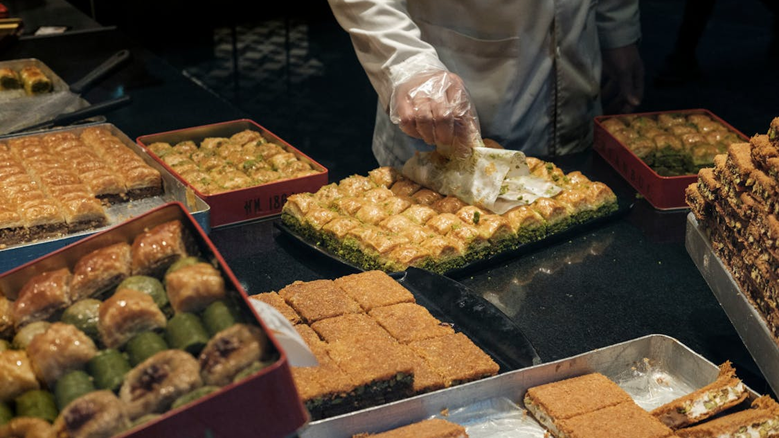A person wearing gloves arranges assorted trays of baklava in a bakery display, with various shapes and toppings visible.